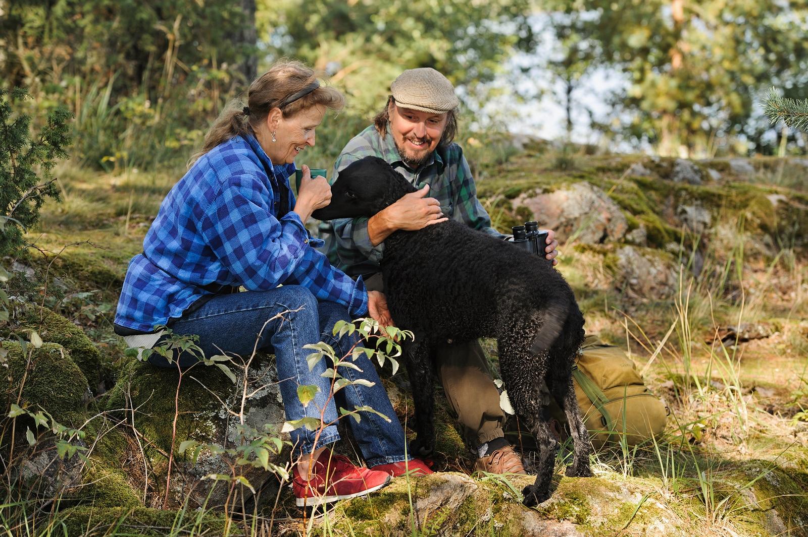 couple-with-dog-in-forest Senior couple playing with their pet dog while out hiking. The dog is a curly haired retriever.