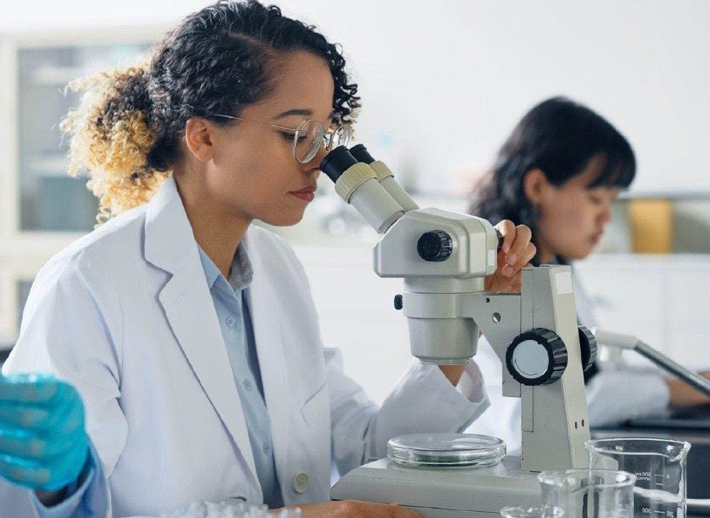明るい研究所で白衣を着用し、顕微鏡を覗き込んでいる女性科学者。
