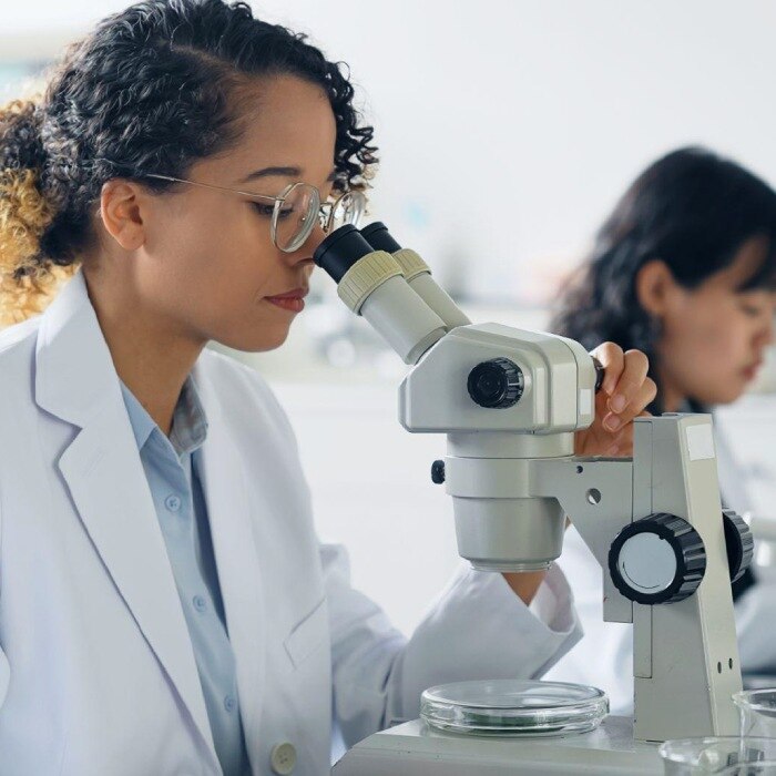 明るい研究所で白衣を着用し、顕微鏡を覗き込んでいる女性科学者。